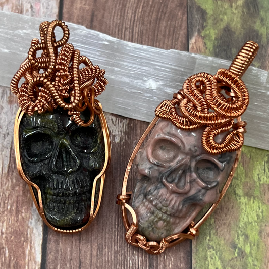 Two skull-shaped pendants with intricate snake-like copper wirework on a wooden surface.